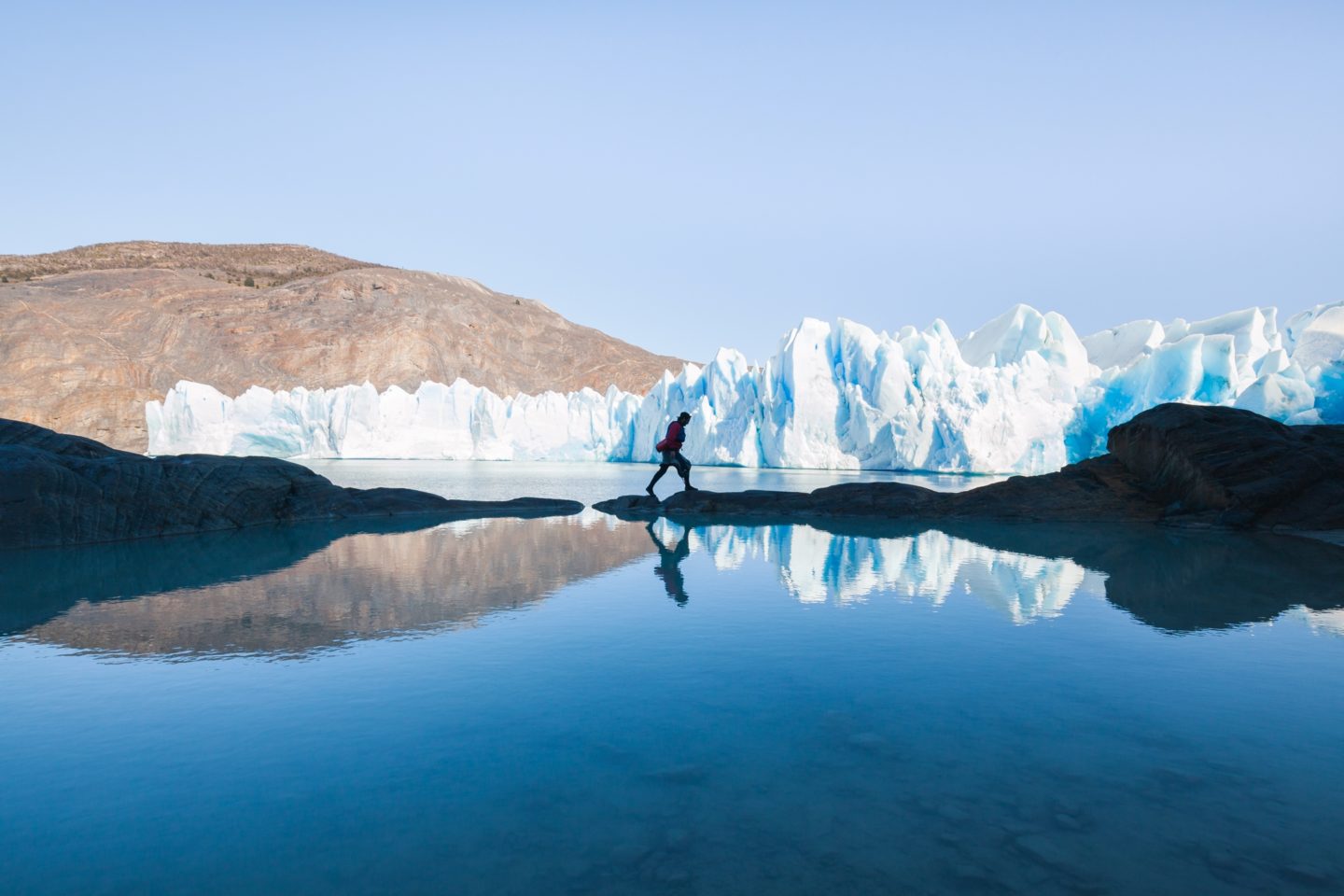 A man strolling along the rocky shoreline with the impressive backdrop of a glacier wall in the distance.