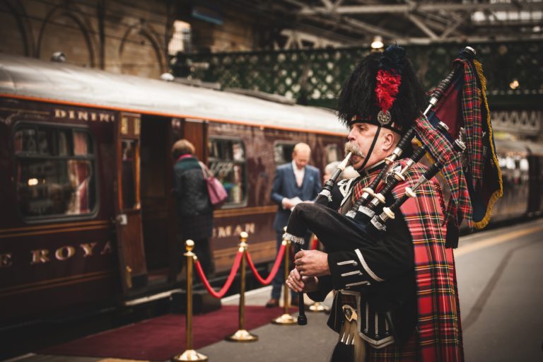 A person in traditional Scottish attire playing the bagpipes beside a luxury train carriage.