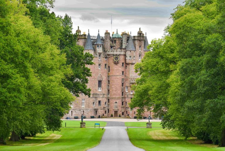 A majestic castle surrounded by lush green trees, with a pathway leading to its entrance.