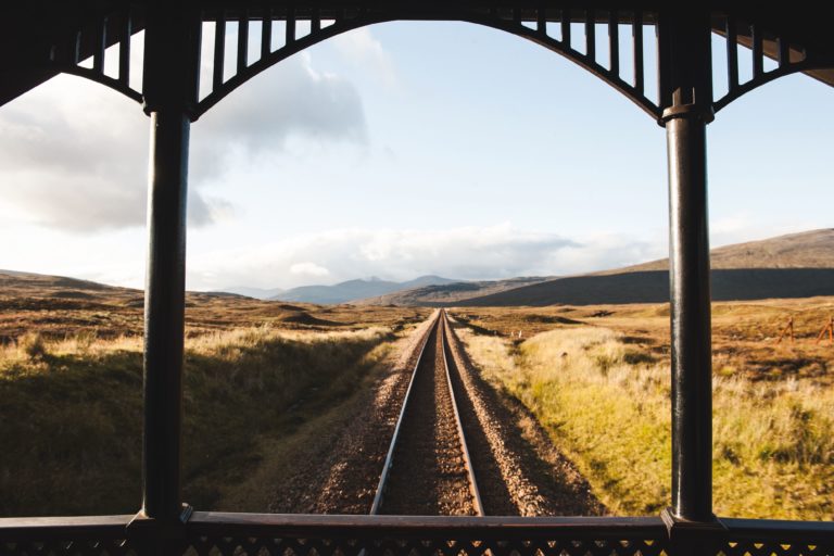 A view from under a metal shelter of a railway track surrounded by open fields and mountains.