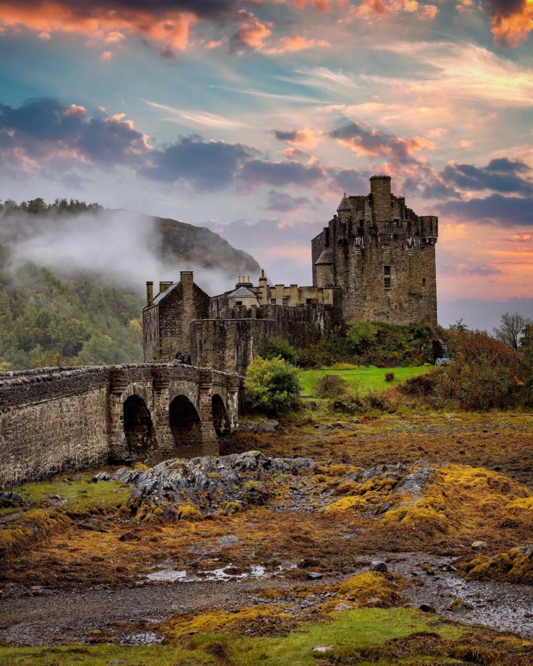 A stone castle and wall on a rocky landscape with greenery.