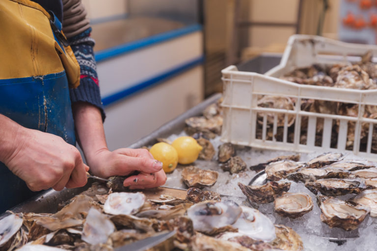 Man shucking oysters over ice at a fish market stall in Carbost, Isle of Skye, with lemon slices nearby.