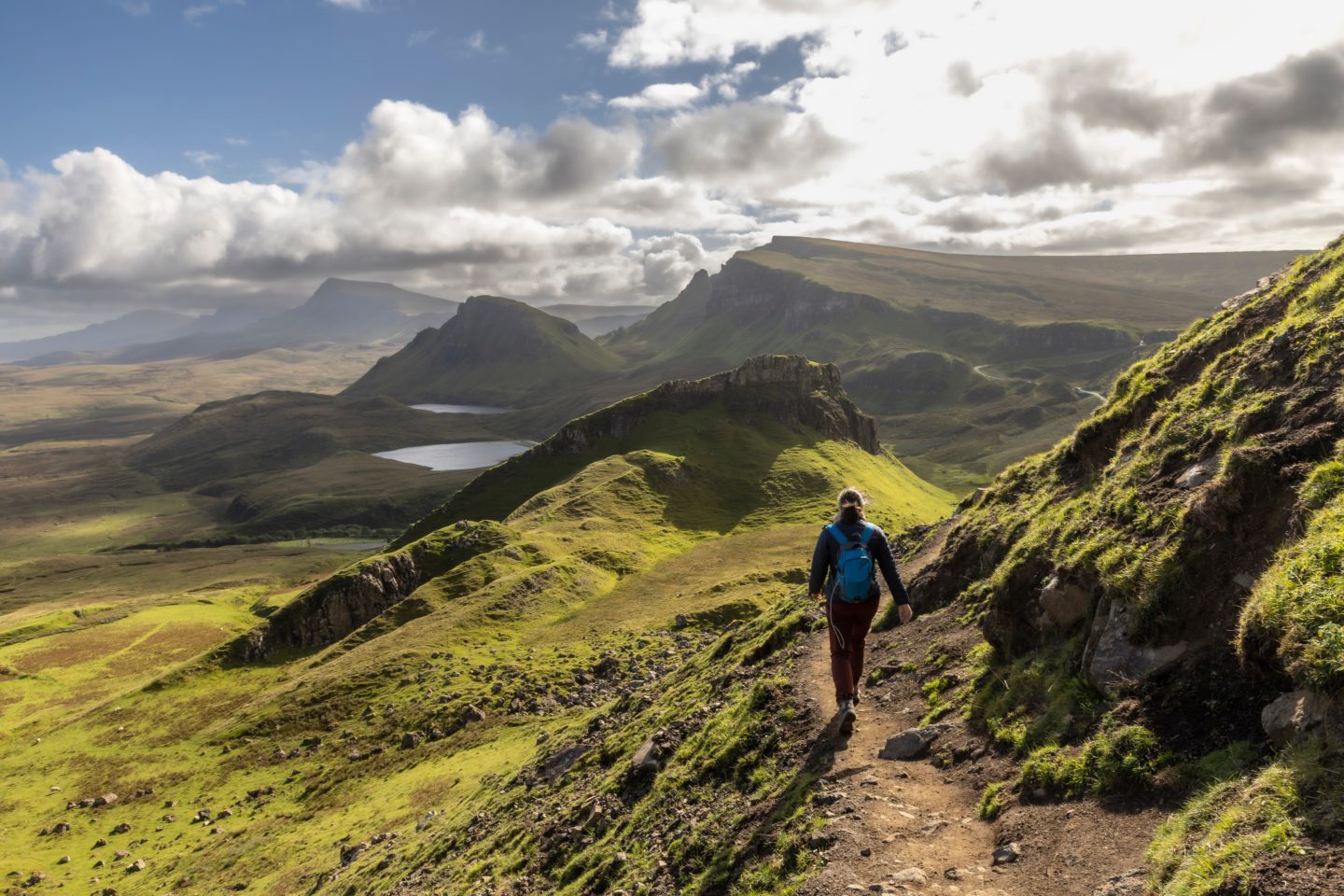 A woman hiking on a scenic trail in the Quiraing, Isle of Skye, surrounded by green mountains and meadows.
