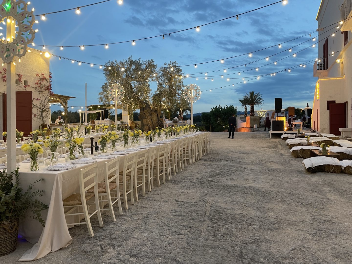 Two long tables under fairy lights on a piazetta are decorated with flowers during a private event in Puglia, Italy.